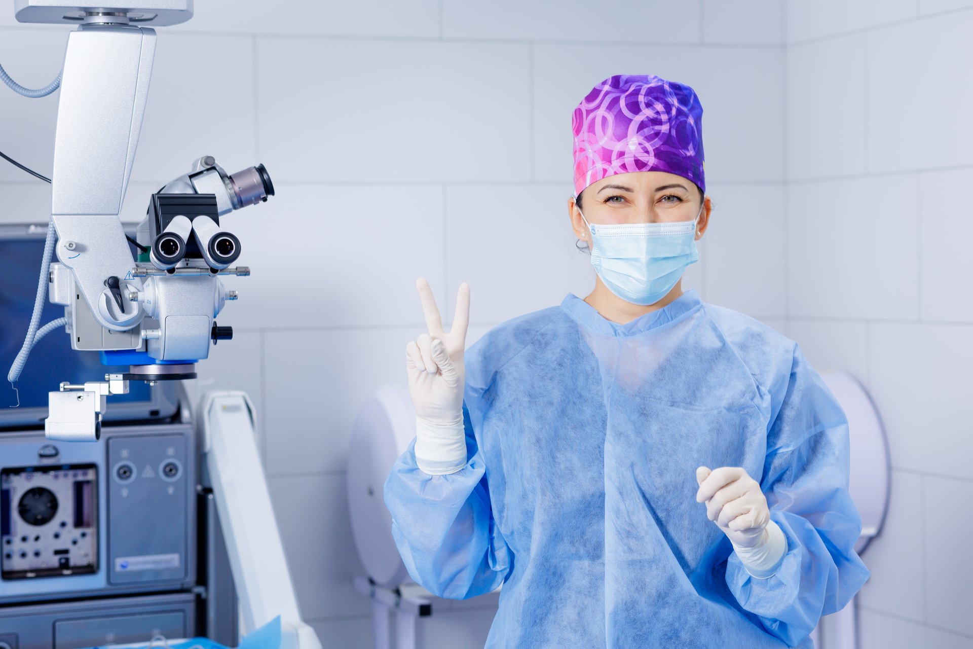 Female eye surgeon in sterile scrubs and mask poses by surgical microscope, signaling readiness and confidence before a precise ophthalmology procedure.