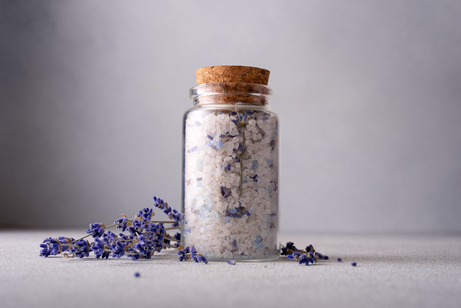 A serene spa setting with a glass jar of aromatic lavender salt sealed with a cork, surrounded by scattered lavender flowers against a grey background, evoking calm and relaxation.
