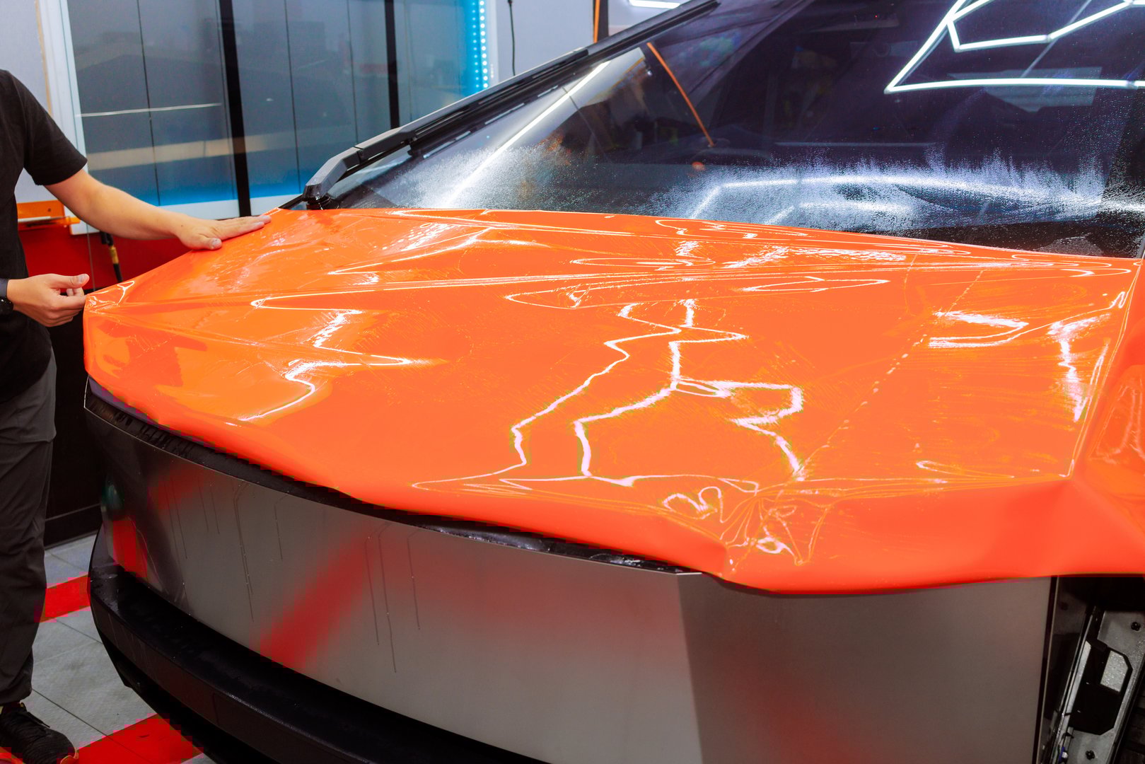 Worker applies bright orange vinyl wrap to car hood at customization workshop in car service
