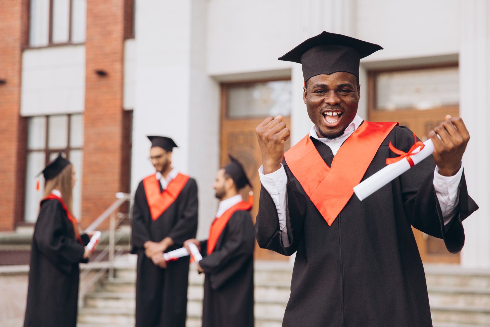A happy African American male graduate show yes and celebrating with friends, wearing traditional cap and gown, holding diploma outside a university building, accomplishment in education.