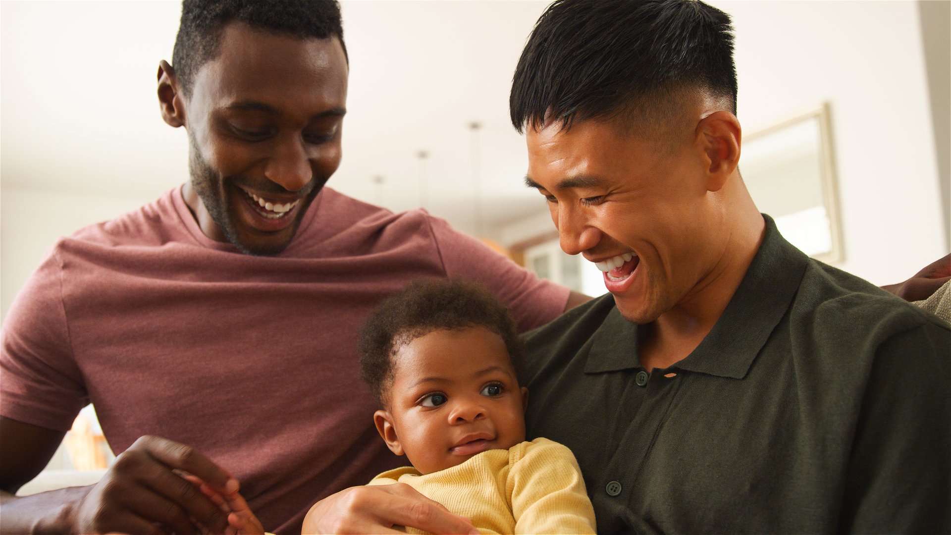 Two men happily interact with their baby in a bright, welcoming home.