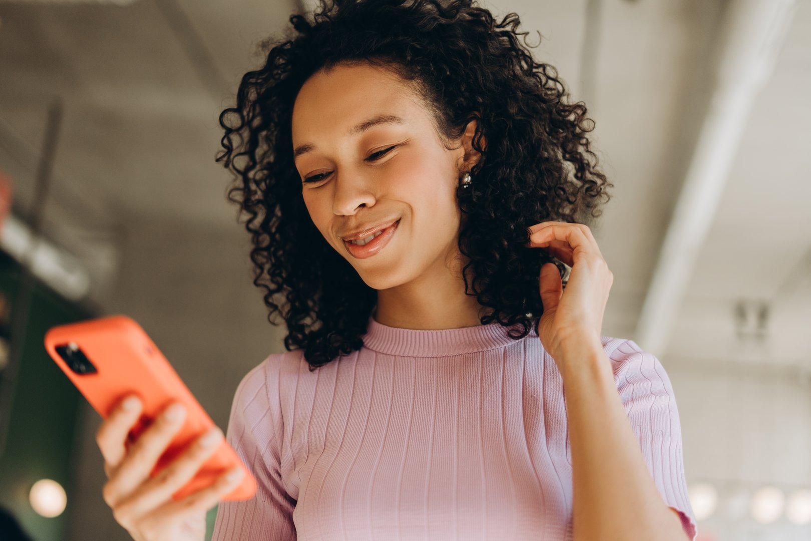 Cheerful young woman with curly hair smiles brightly while holding an orange smartphone and playfully touching her hair, enjoying her time indoors in a modern office setting. Online shopping concept