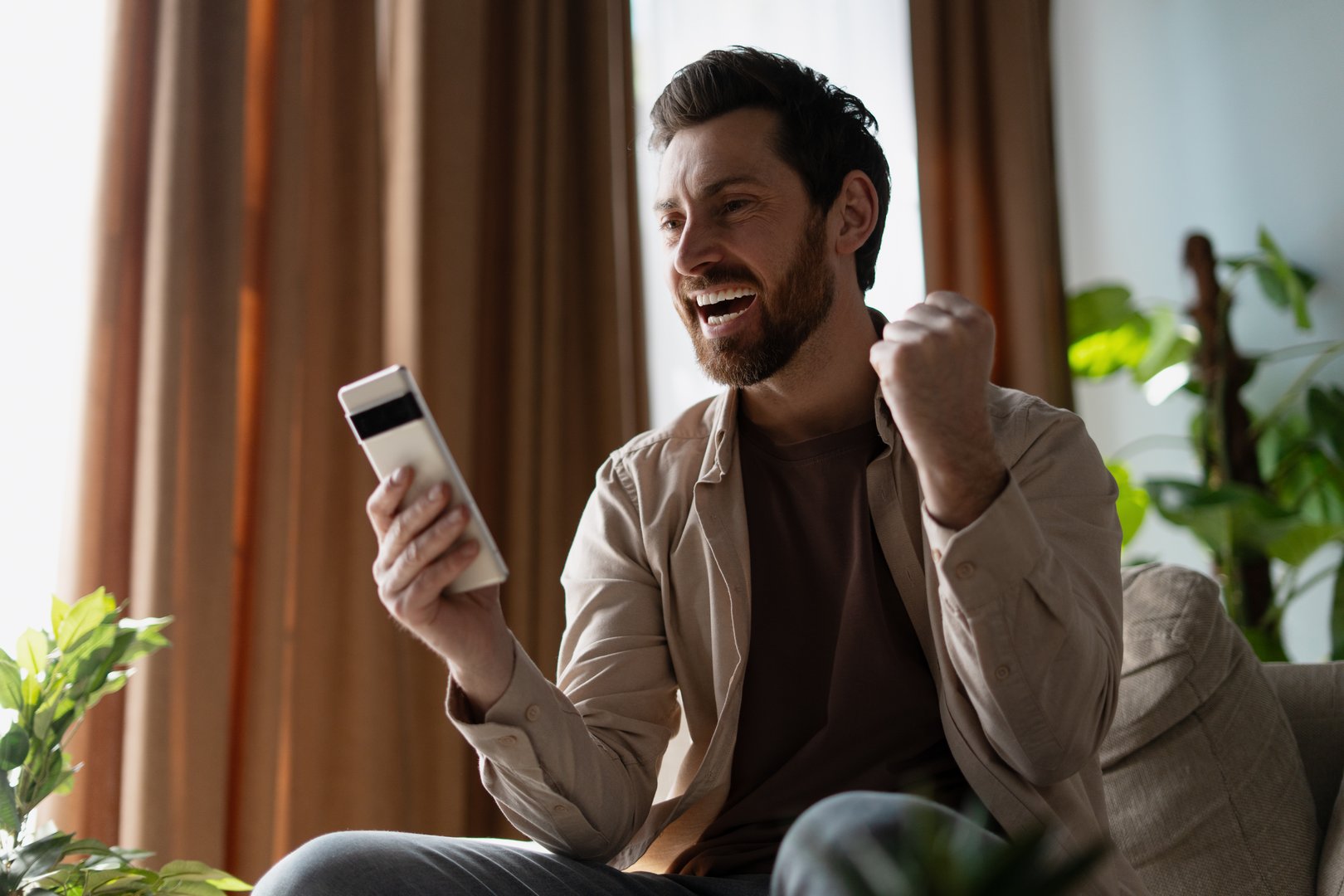 Overjoyed young adult man using mobile phone reading good news, feeling excited about online win or achievement