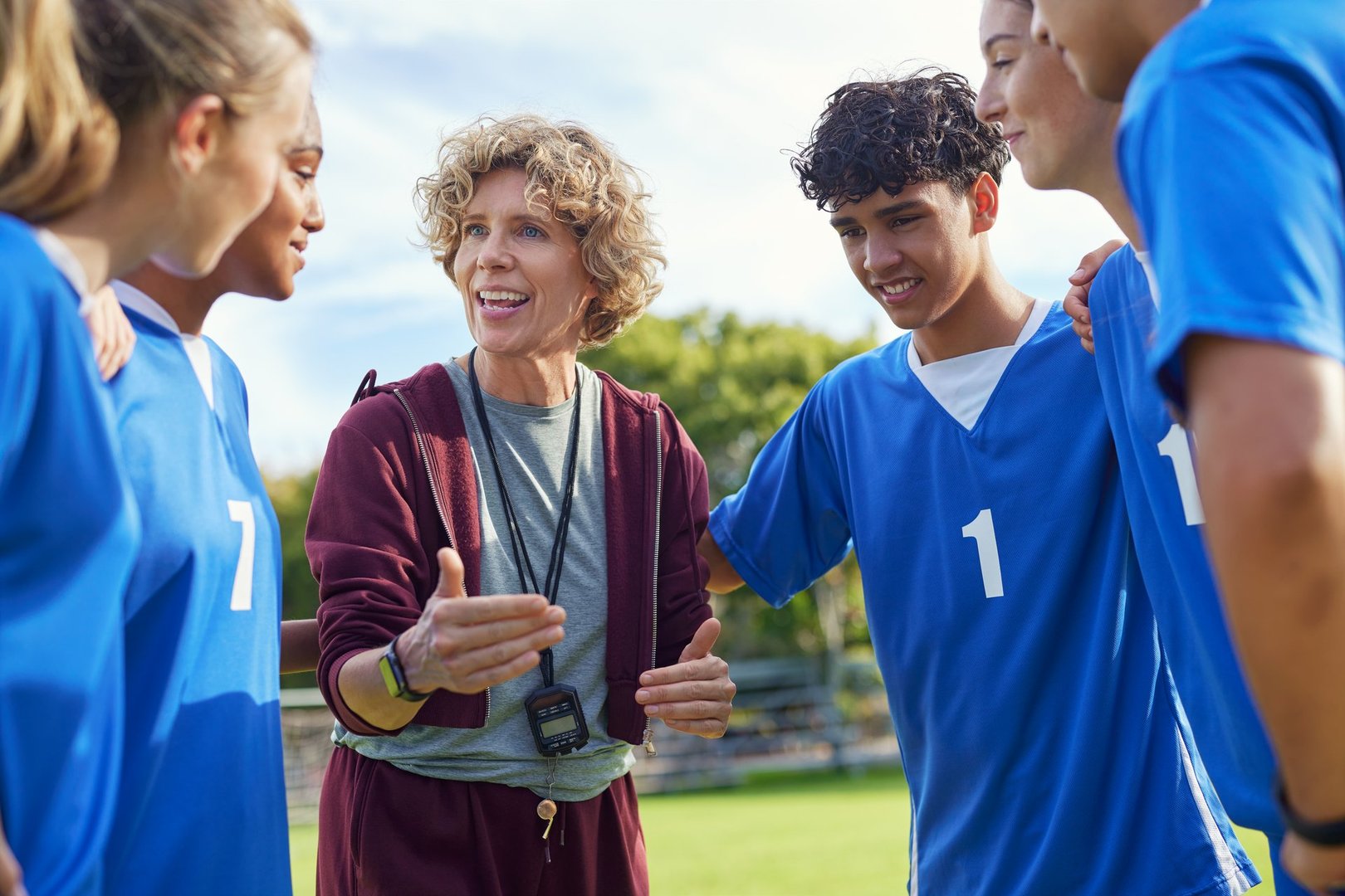 Energetic mature female coach addressing sports team during outdoor practice session. Athletes in blue uniforms listening attentively in a team huddle on the field before match. Coach using hand gesture while speaking and encouraging the team spirit and unity.