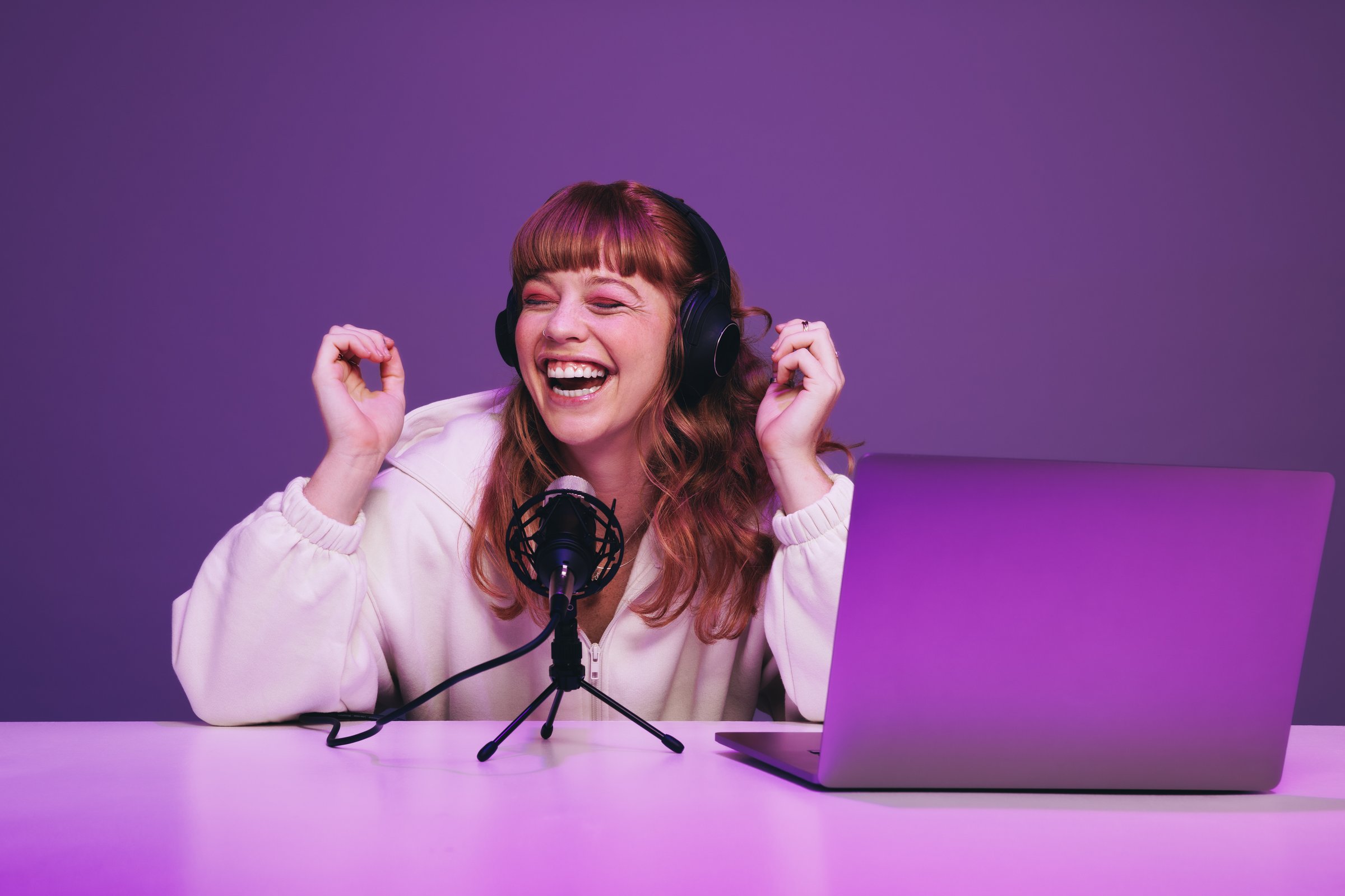 Female podcaster laughing while hosting a live broadcast in a studio. Happy young woman recording an audio show in neon purple light. Woman creating content for her internet podcast.