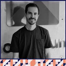 Smiling man in a black t-shirt standing indoors, with logo partially visible in the background.