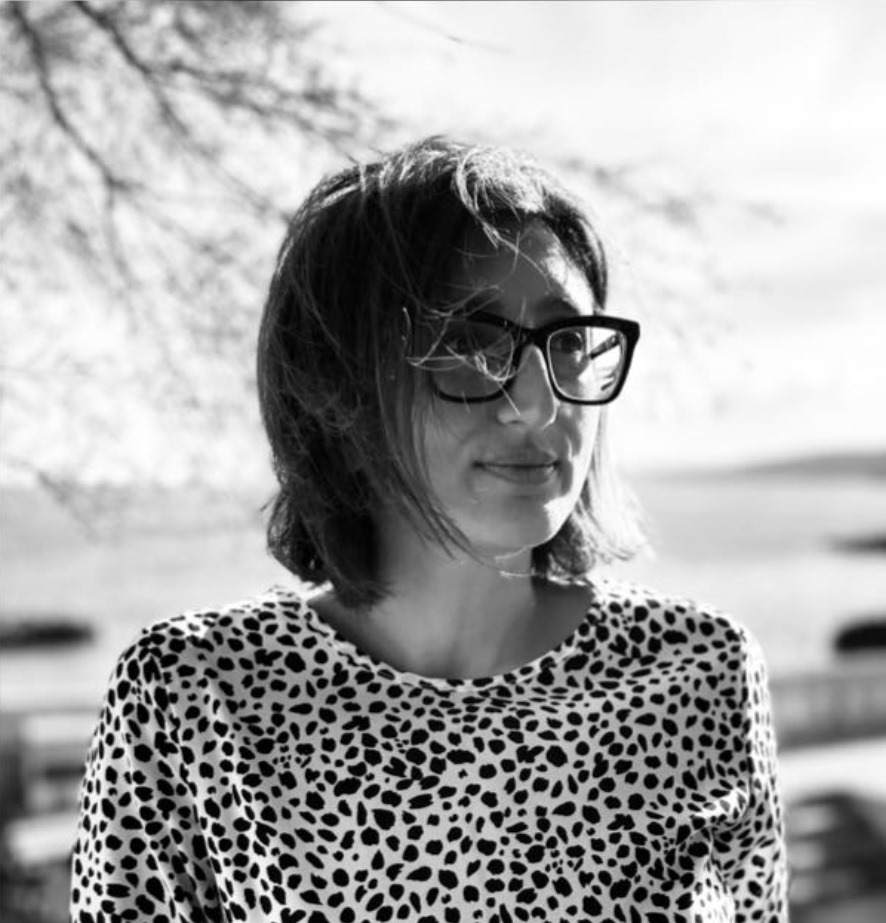 Black and white photo of a person with short hair and glasses wearing a spotted shirt, standing outdoors with blurred background.