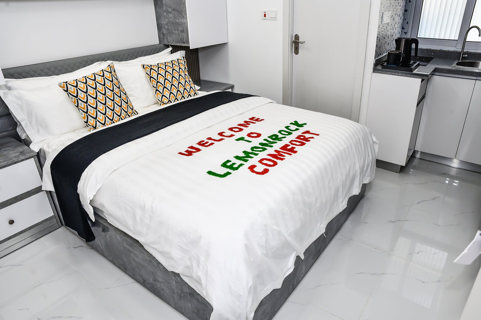 Modern hotel room with a white bed, colorful pillows, and a blanket message reading Welcome to Lemonrock Comfort.