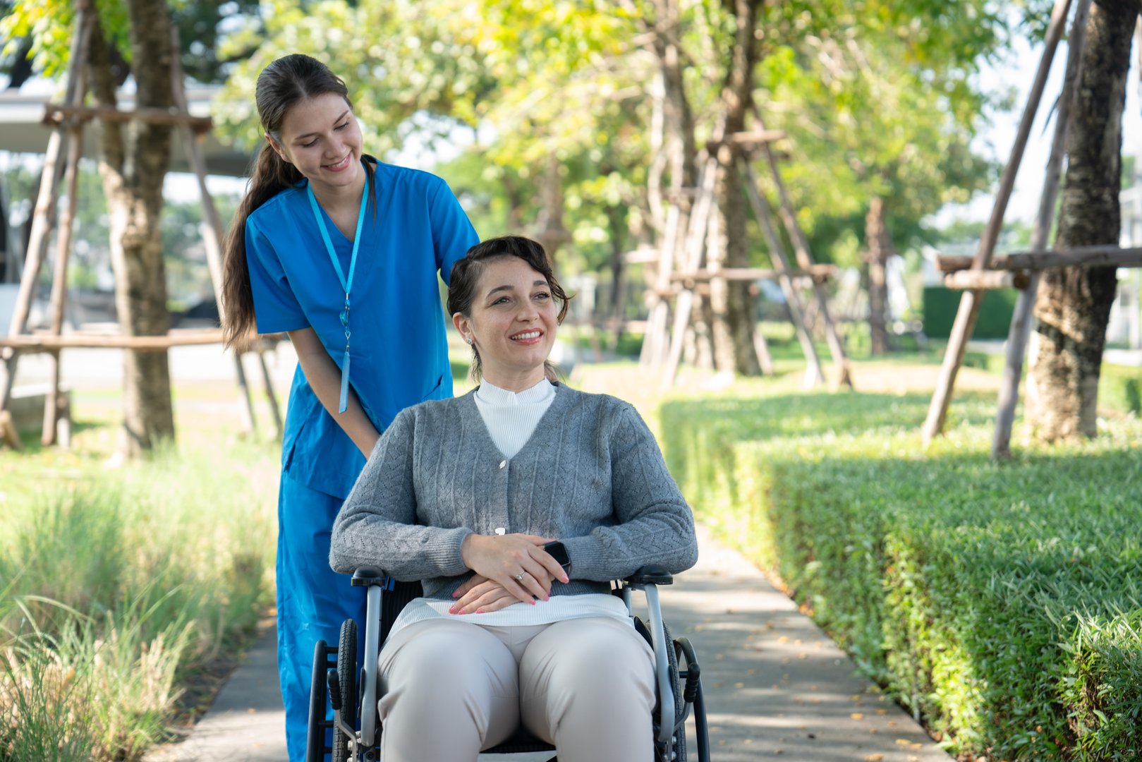 A nurse in blue scrubs smiling and assisting a woman in a wheelchair, enjoying a walk together in a sunny park.