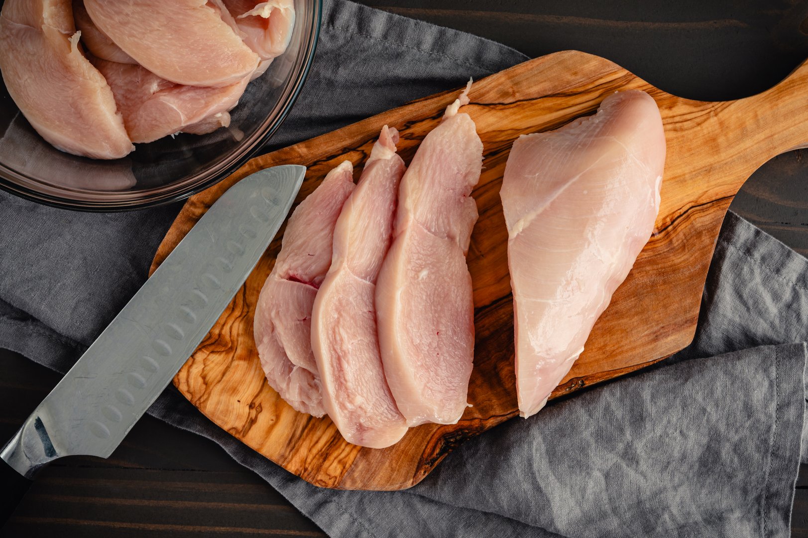 Raw chicken breast slices on a wooden cutting board with a chef's knife