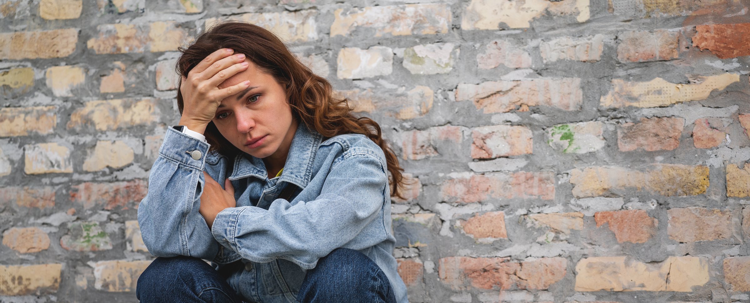 Overwhelmed and depressed urban woman sits next to brick wall outdoors in the city with hand on her forehead. Stress and mental illness.