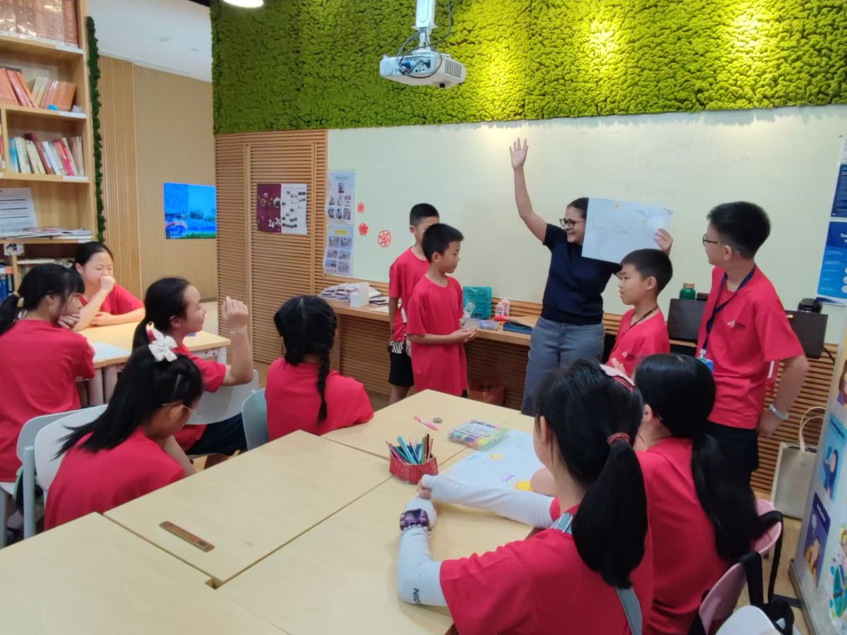 Students in red shirts sit at desks while a teacher leads a classroom activity with a projector overhead.