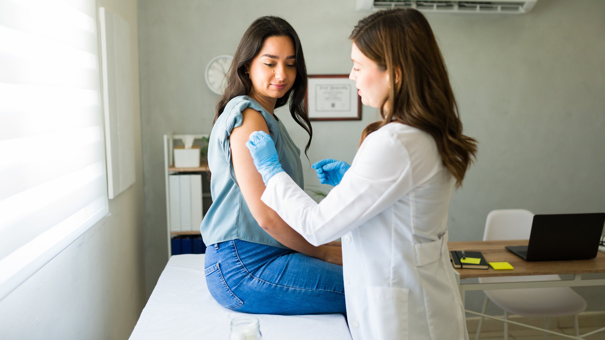 Young Hispanic woman is getting vaccinated by a professional medical worker wearing gloves in a doctor's office