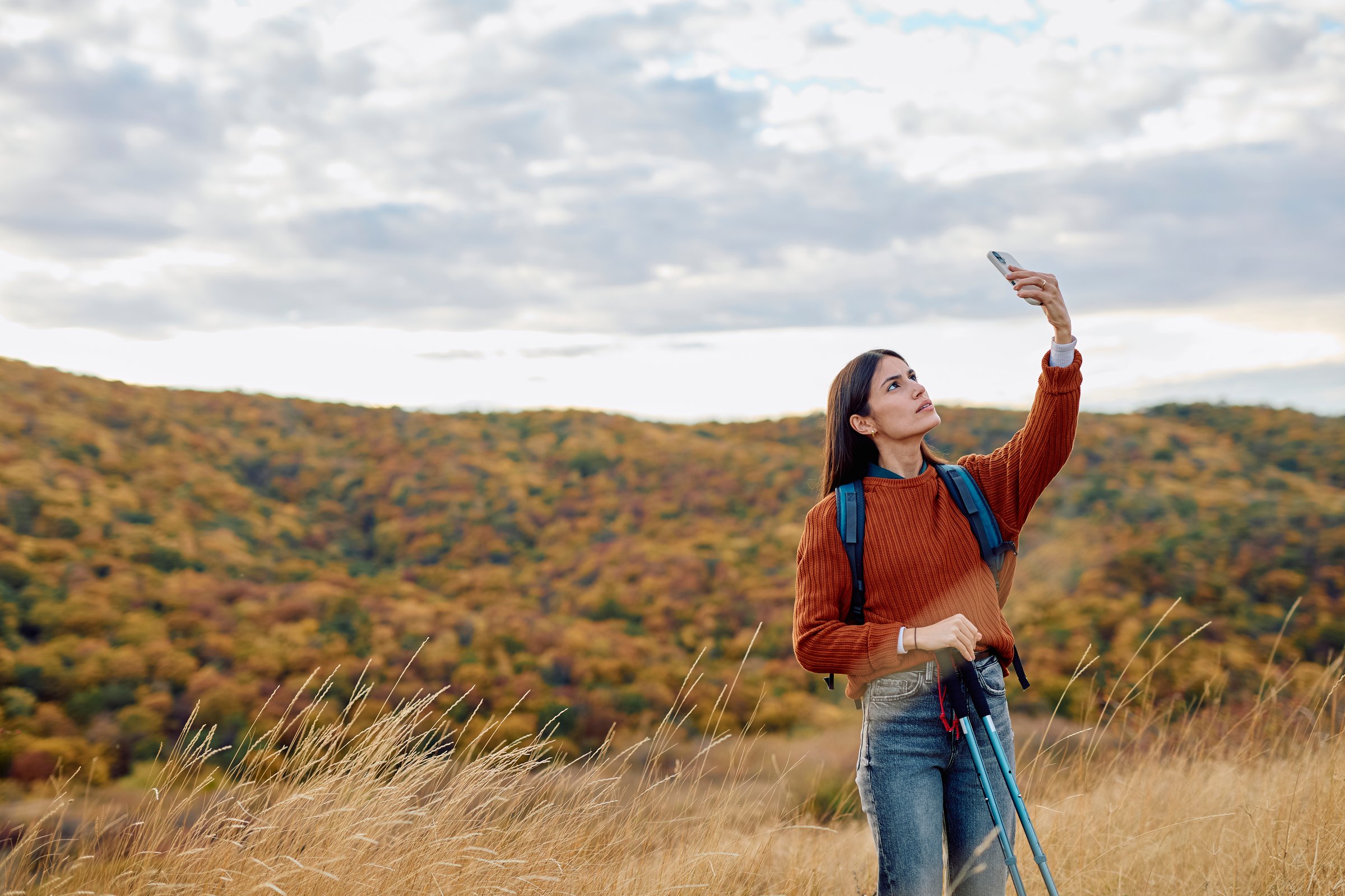 Young woman standing on a hill in autumn nature, holding up a smartphone, searching for a cellular mobile signal