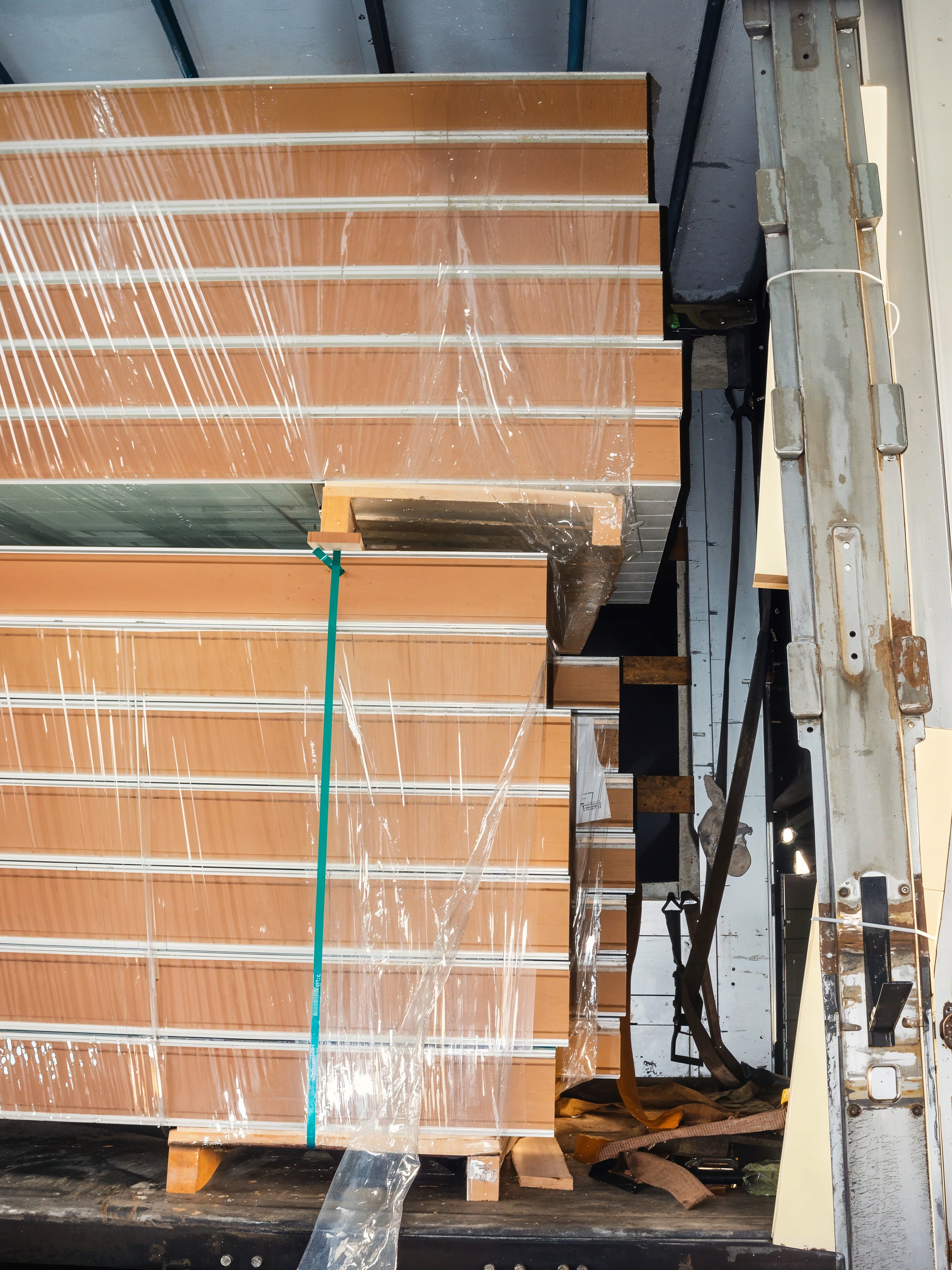 Stacked panels wrapped in plastic inside a warehouse or transport vehicle. Wooden pallets and other equipment are also visible.