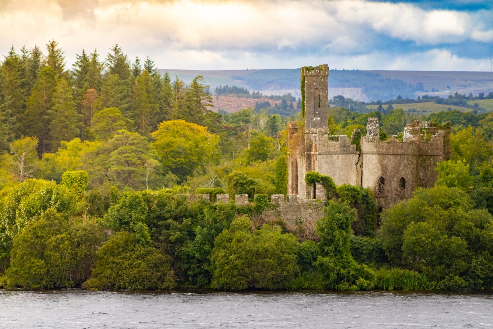 A scenic view of McDermott's Castle in ruins, nestled among dense woodland on Castle Island in Lough Key, County Roscommon, Ireland. The structure is surrounded by lush green trees with hills in the background