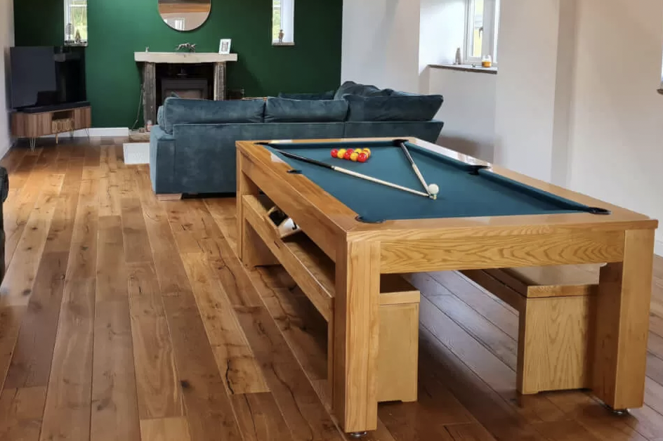 A pool table and benches in a living room with a green wall, sofa, wooden floor, TV, and mirror above a fireplace.