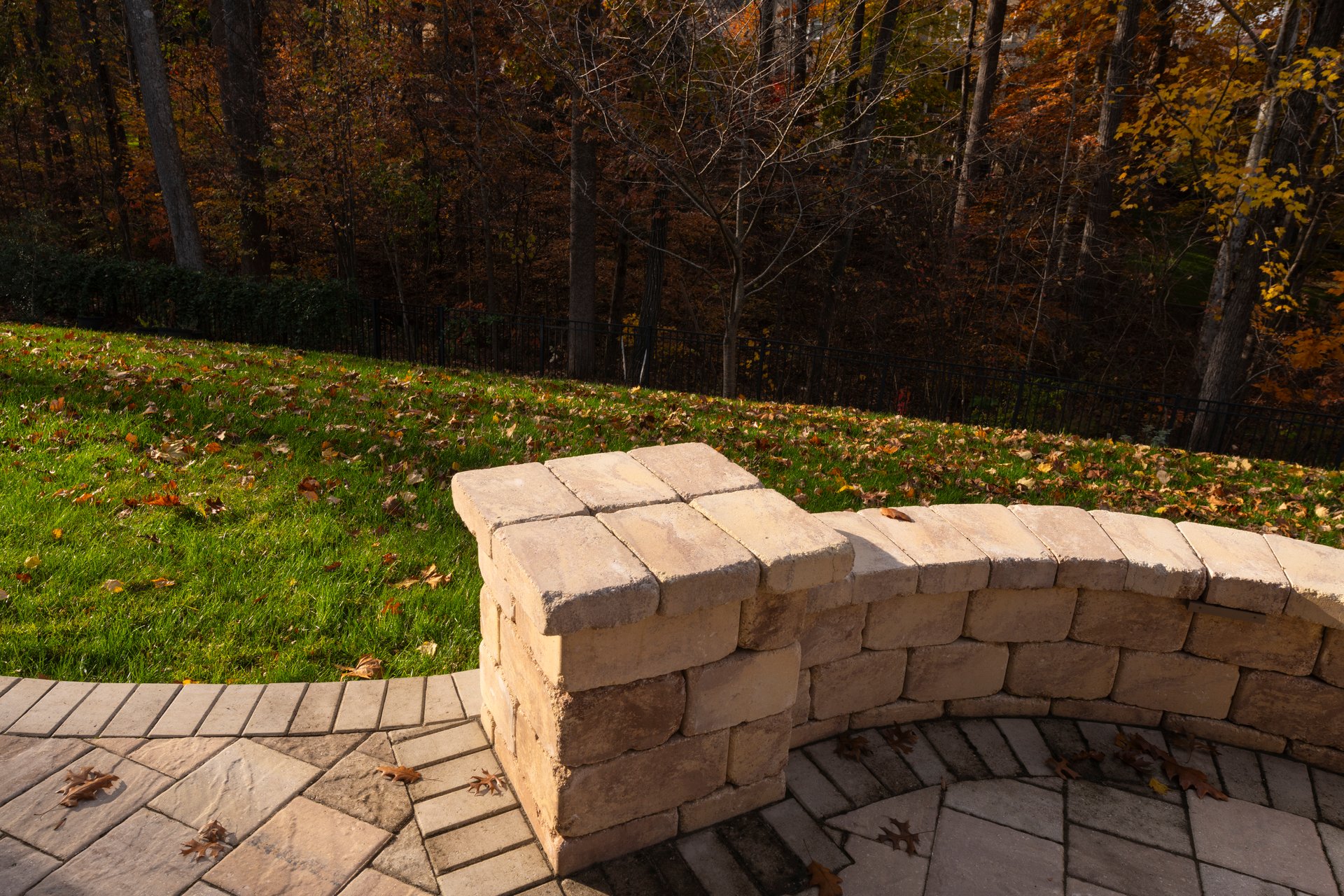Close-up of a curved stone patio wall surrounded by a grassy backyard covered with autumn leaves, offering a warm and inviting outdoor atmosphere.