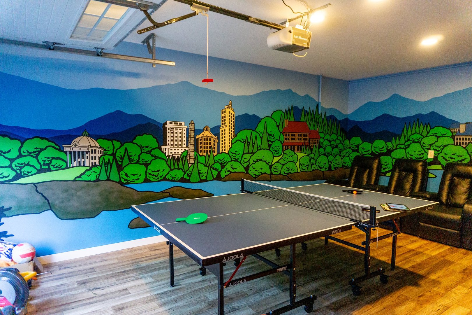 Ping pong table in room with a vibrant mural of a cityscape and mountains, featuring reclining chairs and wood flooring.
