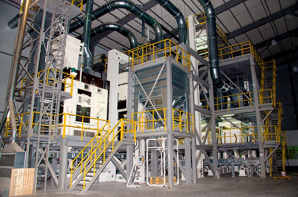 Industrial plant interior with large machinery, metal platforms, stairs, and ventilation pipes in a spacious warehouse.