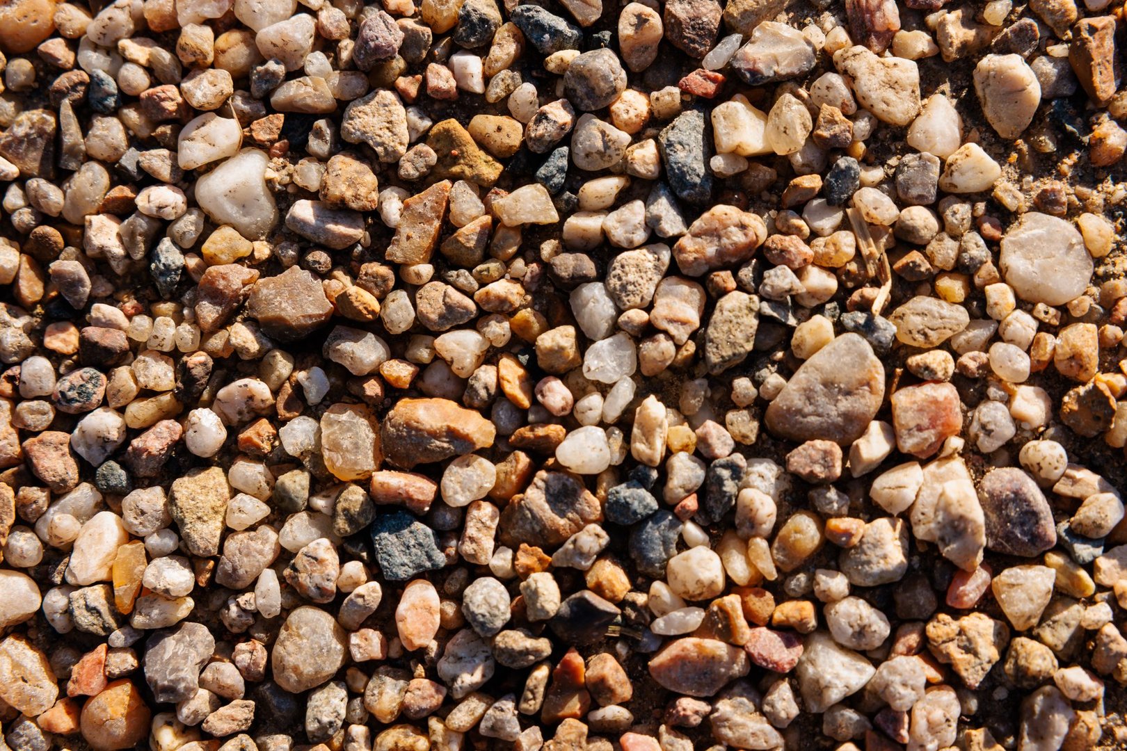 Close up macro view of colorful river pebbles forming a natural gravel surface. Warm sunlight reveals the texture of rounded stones, quartz fragments and mineral details. Rich earth tones and varied shapes create a vibrant abstract background ideal for construction or landscaping visuals, eco concepts, beach themes, web design, packaging, posters, signage and large format prints in high resolution.