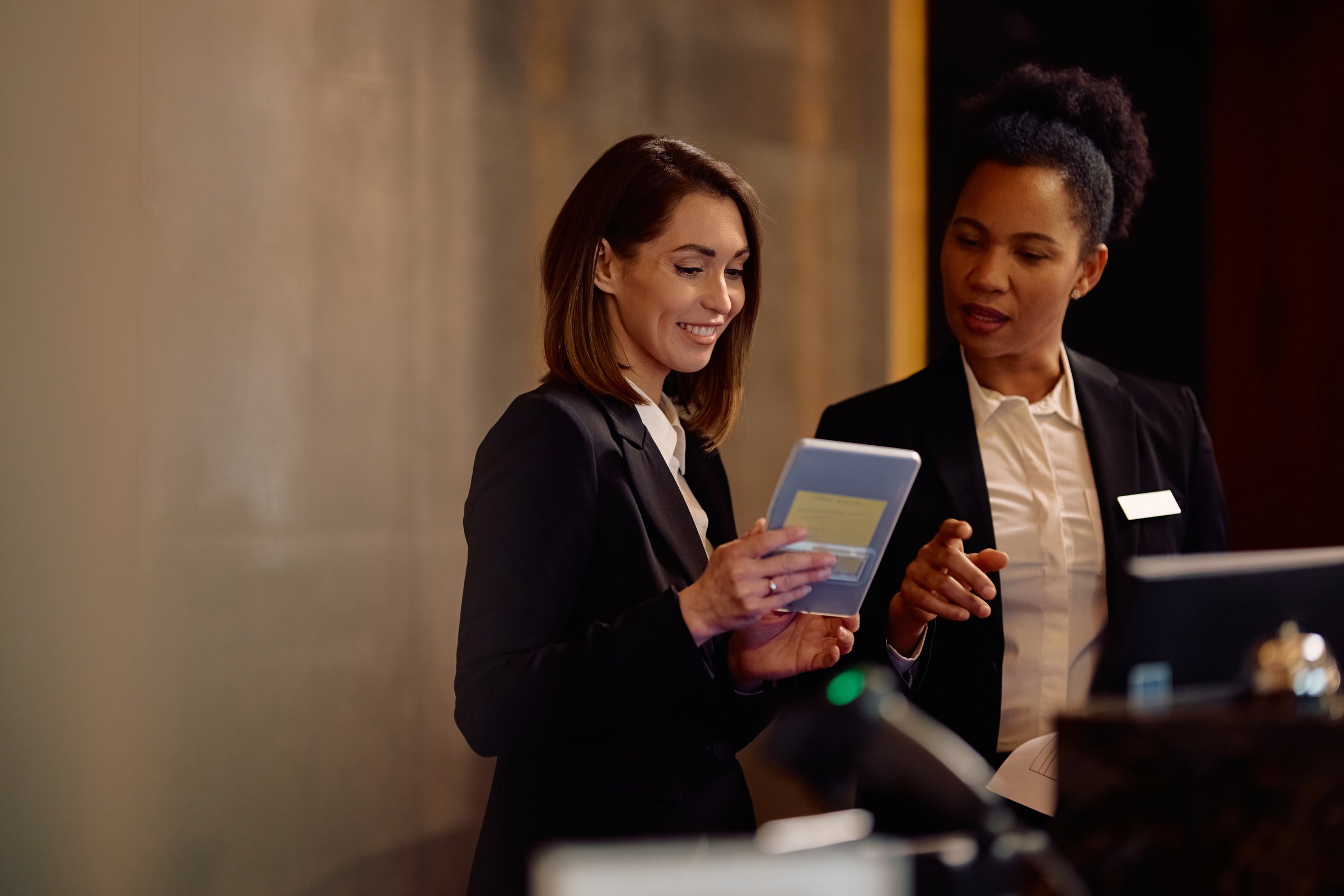 Happy hotel manager and her African American colleague working on touchpad at reception desk.