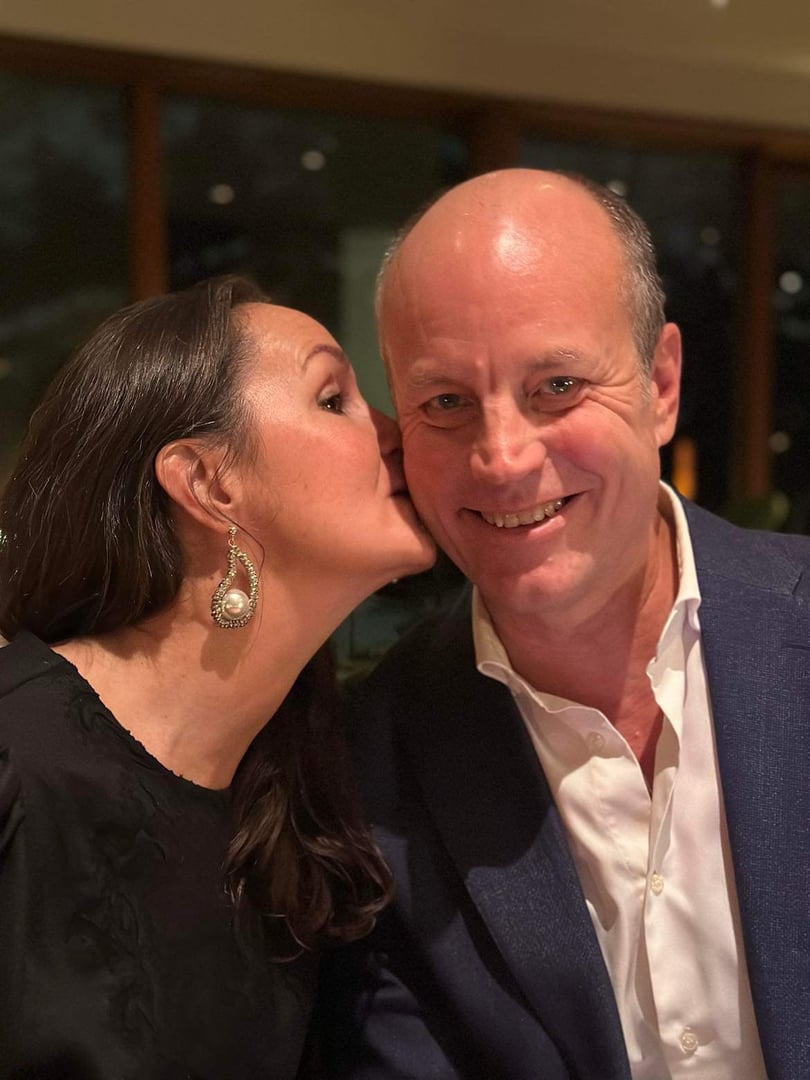 A woman whispers to a smiling man at a dining table. She wears a pearl earring. Both appear happy in a cozy setting.