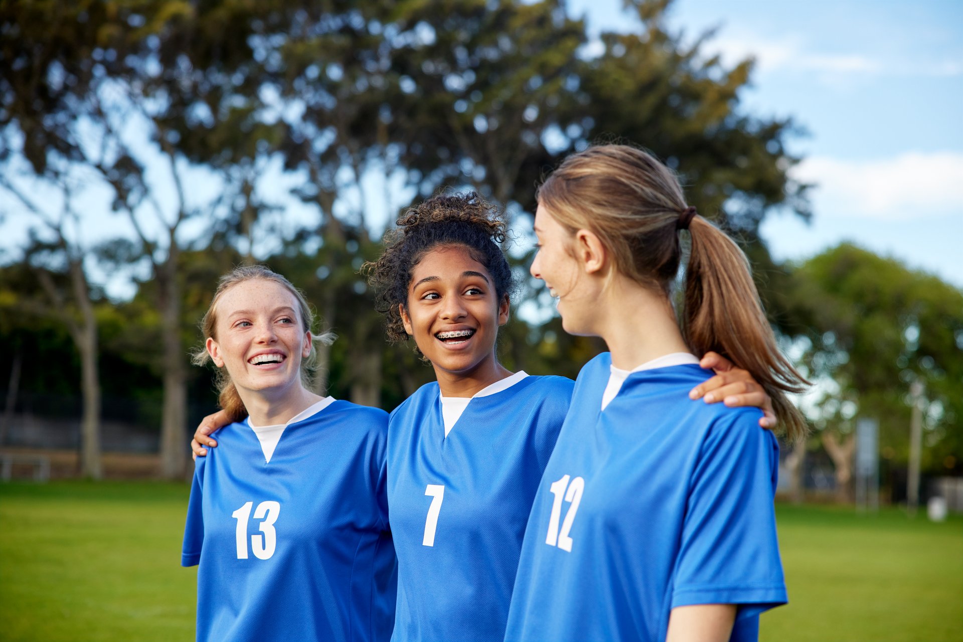 Three smiling athletic girl in blue uniform walking together on grassy field after game. Friendly sports female players sharing a moment of laughter and support. Confident teenager team walking arm in arm and enjoying post game conversation.