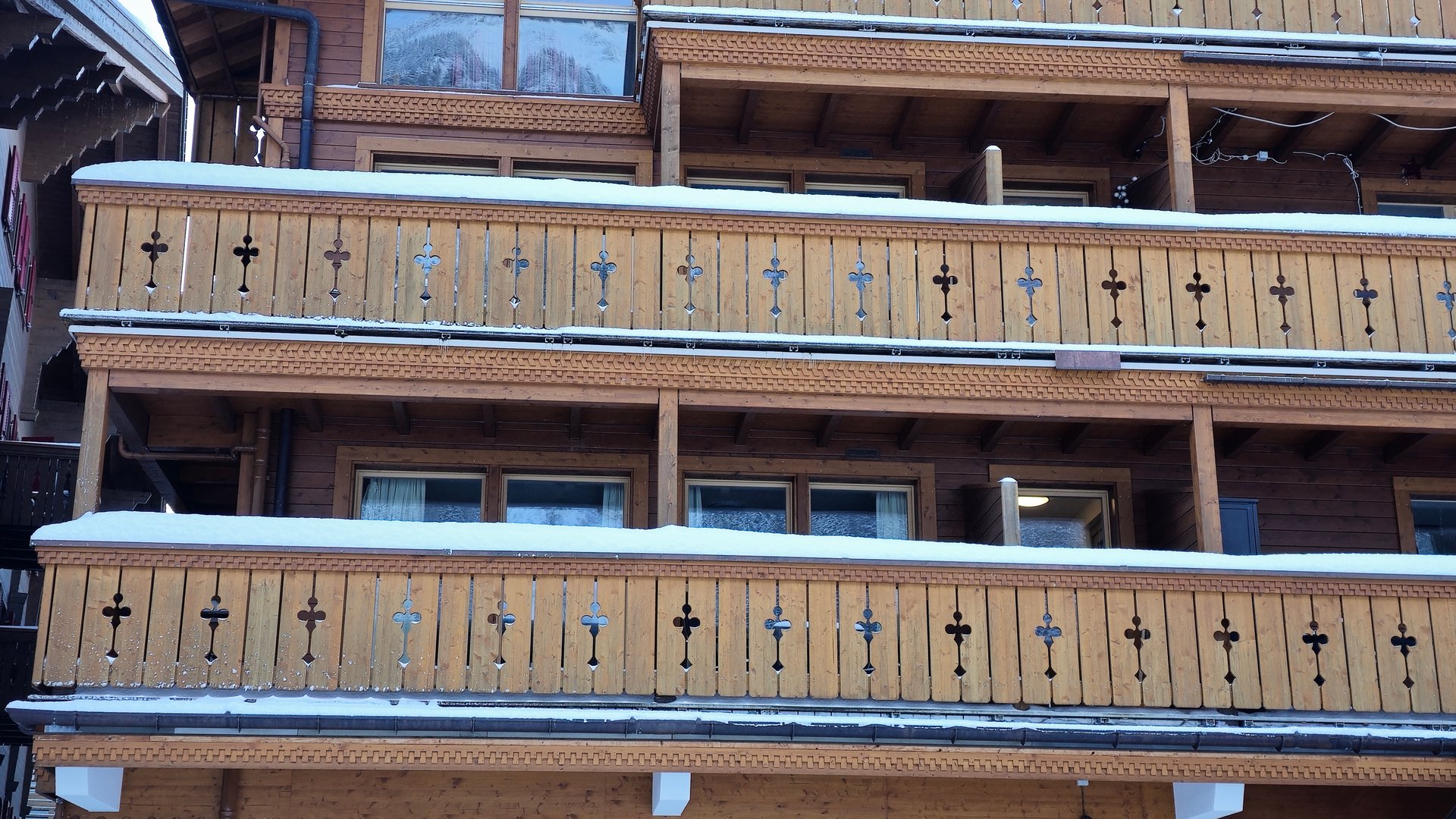A traditional wooden chalet with multiple balconies adorned with decorative railings. Snow rests on the railings and roof, indicating a winter setting in a mountainous or alpine region.