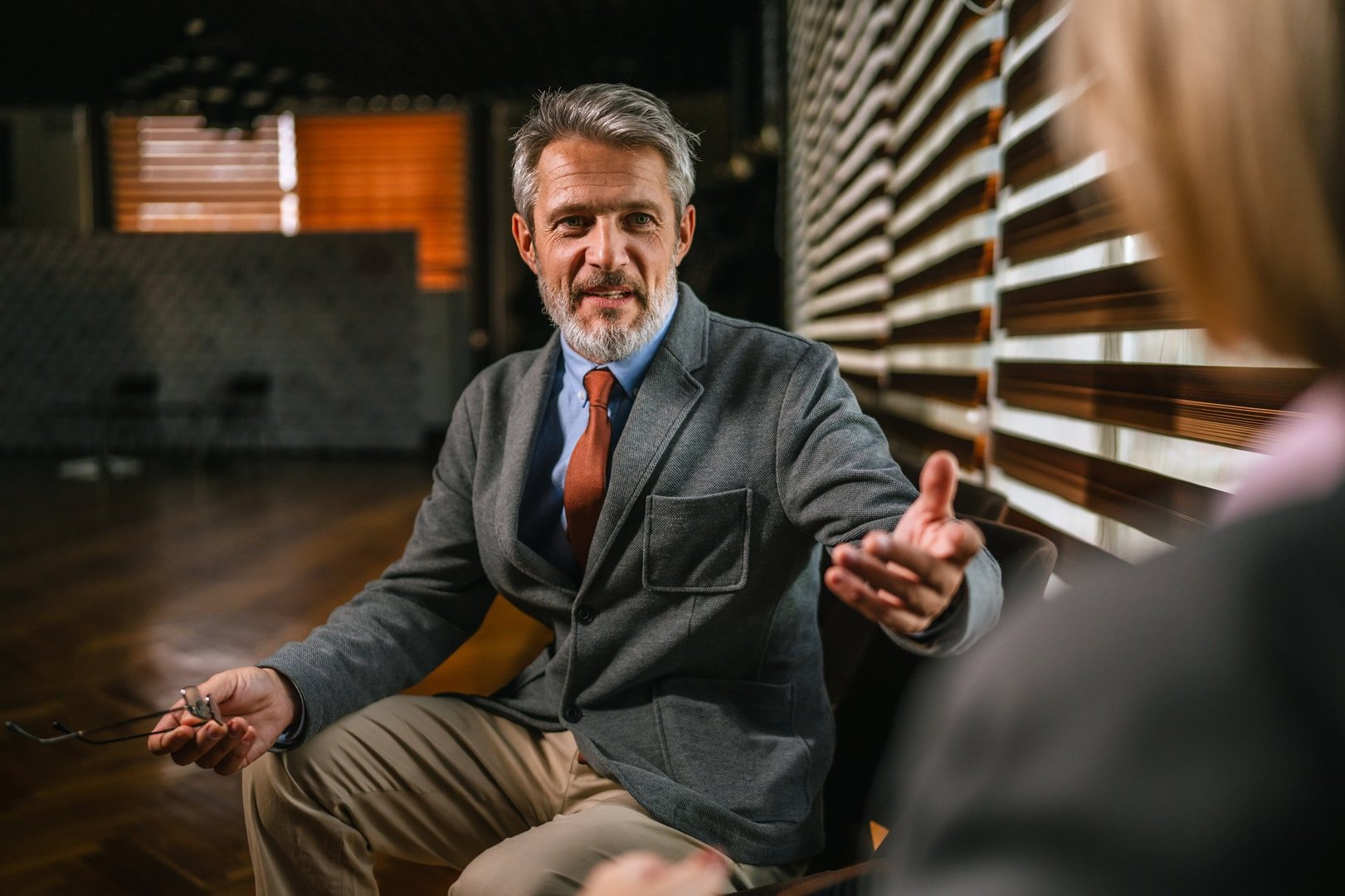 Mature businessman speaking and gesturing with hand, holding eyeglasses while talking to an unseen colleague during an informal business discussion or consultation in a modern office space