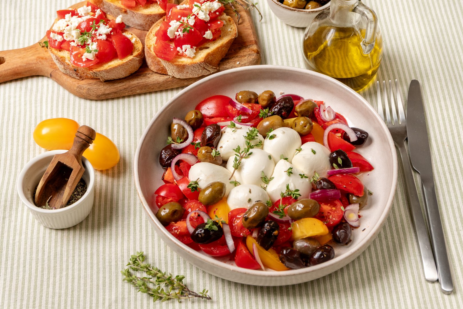 salad with colorful tomatoes, mozzarella balls, and a variety of olives, garnished with herbs, served in a bowl on a textured tablecloth with olive oil and bread