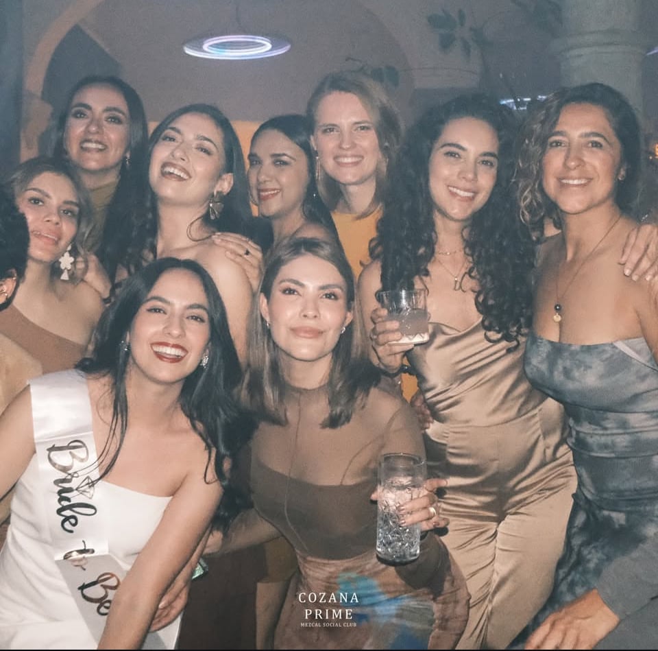 A group of women smiling and posing together at a party, with one wearing a "Bride to Be" sash. Indoor setting.