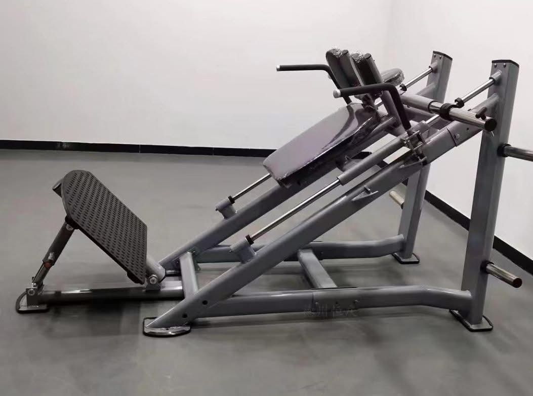 A leg press machine in a gym with a black footplate, padded seat, and metal frame, set against a plain gray floor and white wall.