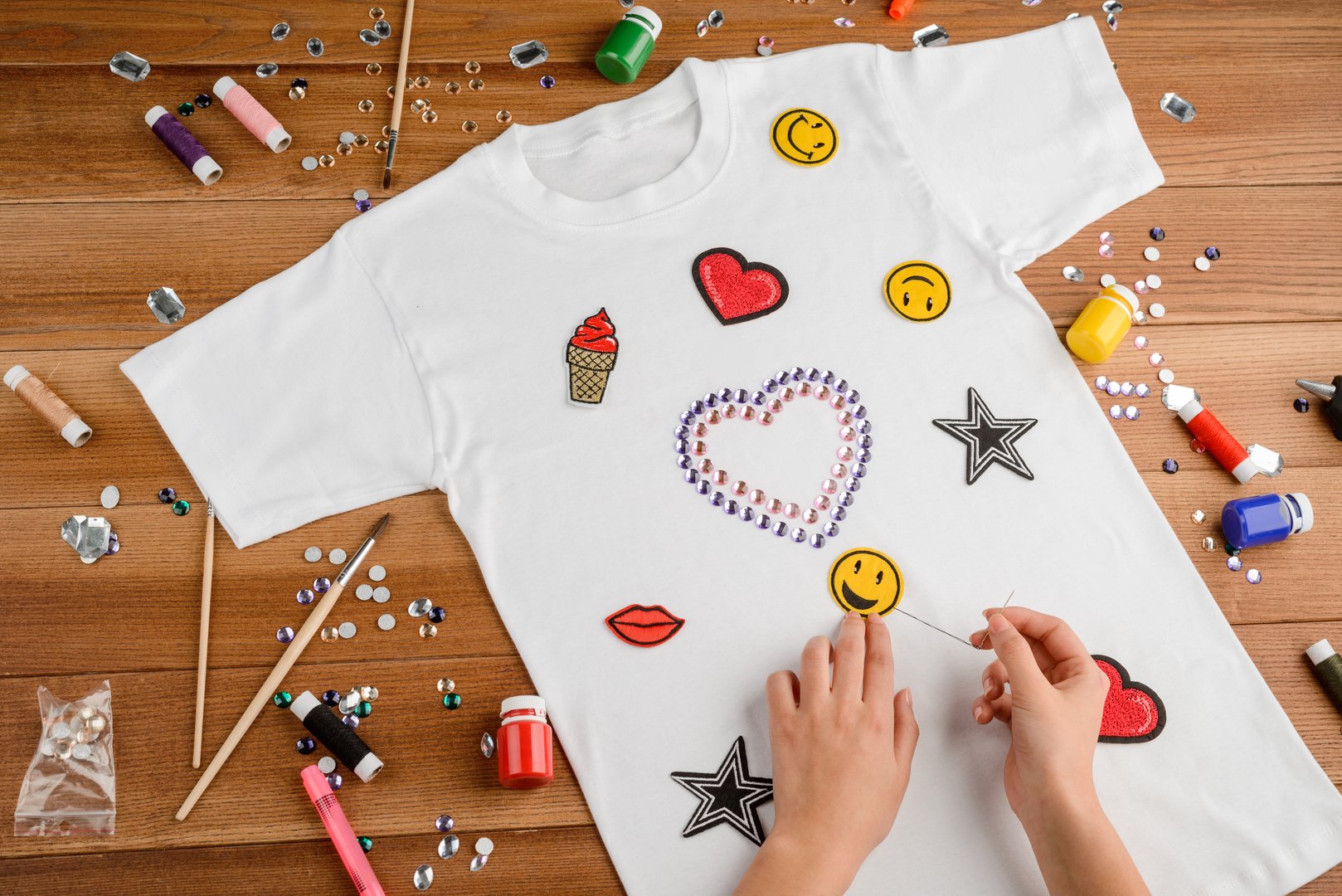 Girl applying patches and rhinestones to a white t-shirt on a wooden craft table. Handmade design for clothes.