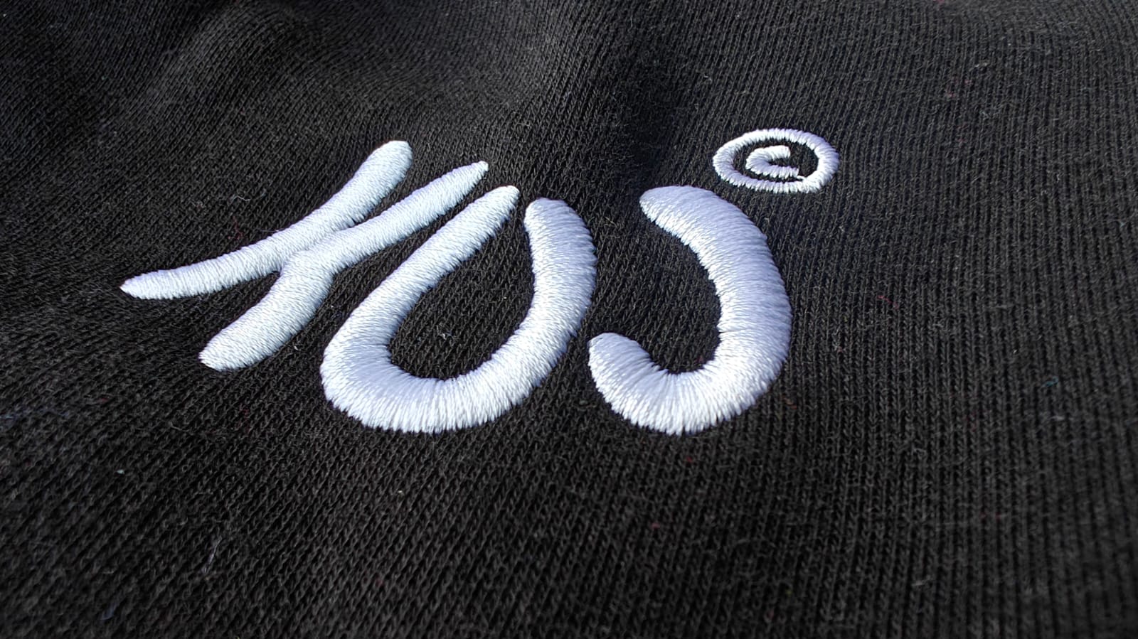 Close-up of white embroidered logo on black fabric, featuring artistic lettering with the letter C in a circle.