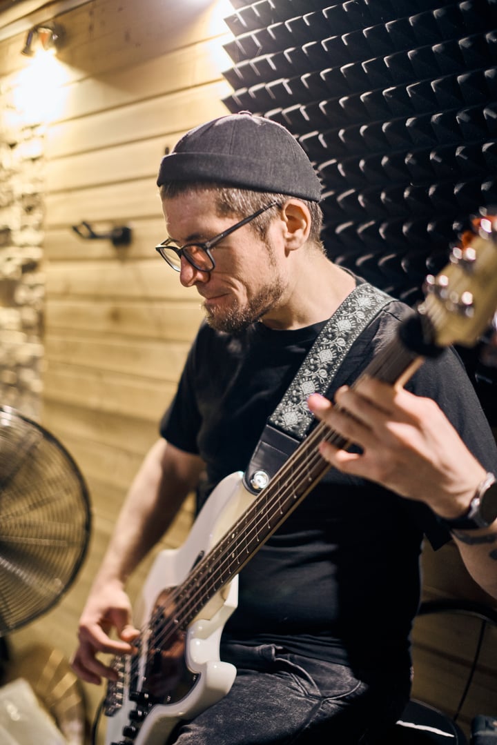 Young adult male with short hair and glasses plays bass guitar passionately in wooden studio. Warm lighting highlights focused expression. Acoustic panels and fan visible