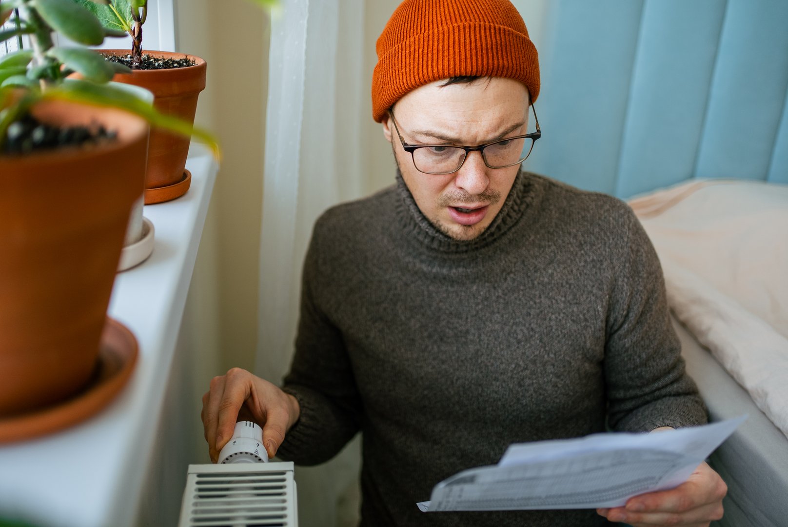 Man in orange hat and warm sweater adjusting a radiator thermostat, looking shocked and worried, holding a huge utility bill showing increasing living costs during an energy crisis in winter