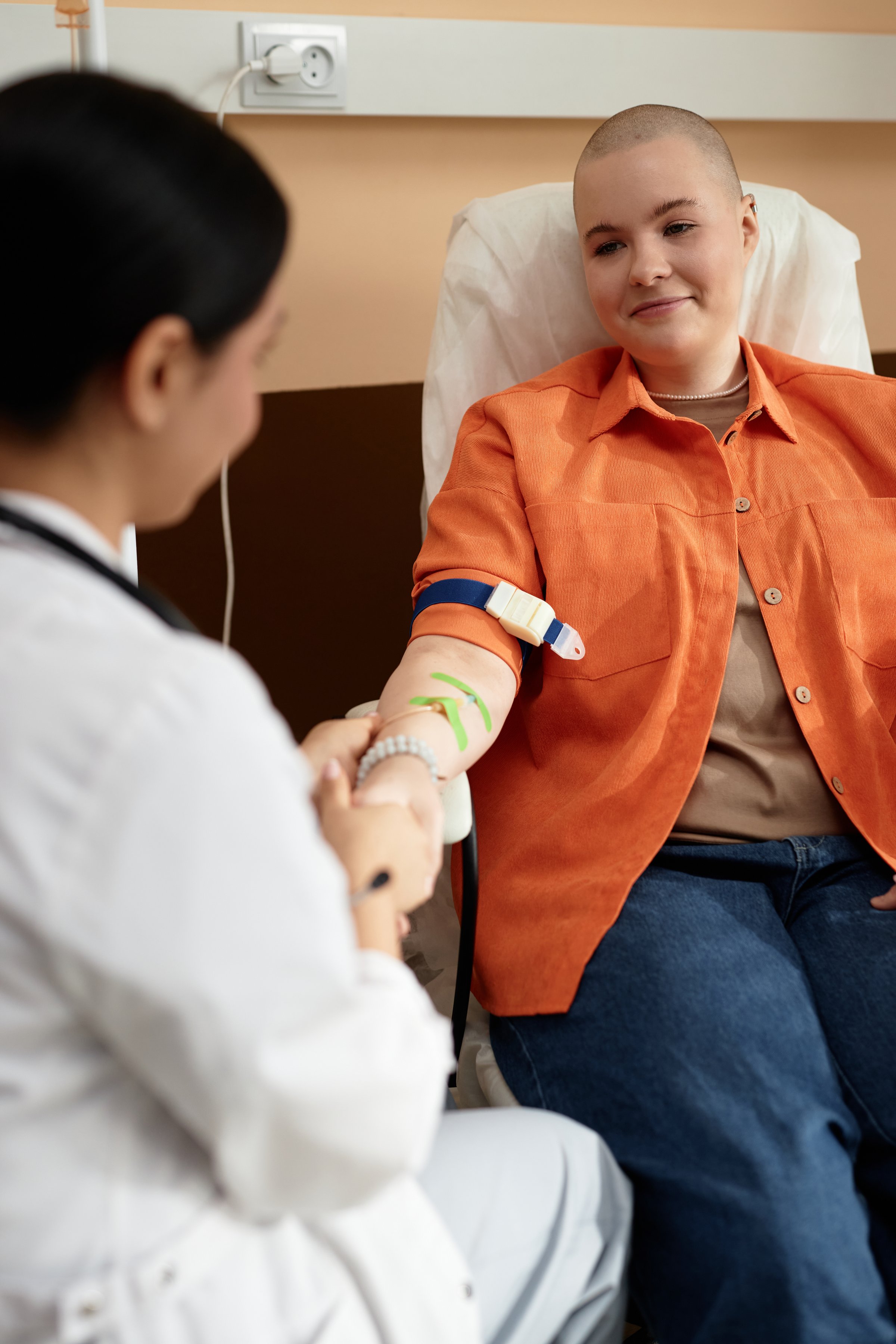 Vertical portrait of smiling bald woman receiving chemotherapy treatment in procedure room and holding hands with caring nurse