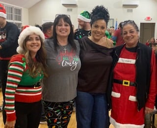 Four women in festive attire posing together at a holiday event, with one wearing a Santa suit and others in Christmas-themed clothes.