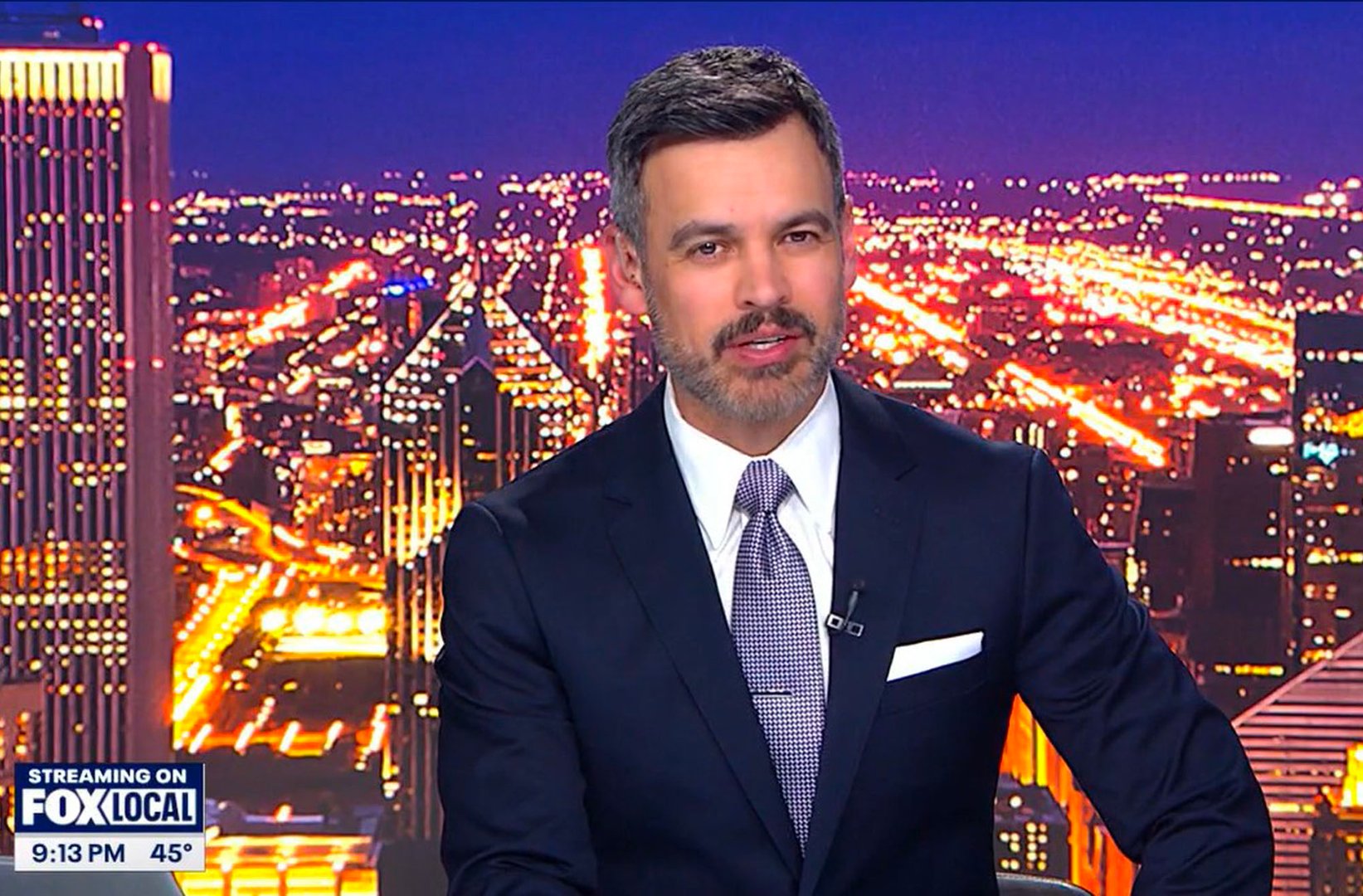 News anchor in a studio with a cityscape backdrop, displaying time, temperature, and network details on screen.
