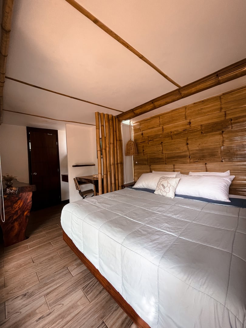 Cozy bedroom with bamboo wall, king-size bed, white linens, and wooden accents. Natural light from a nearby window.