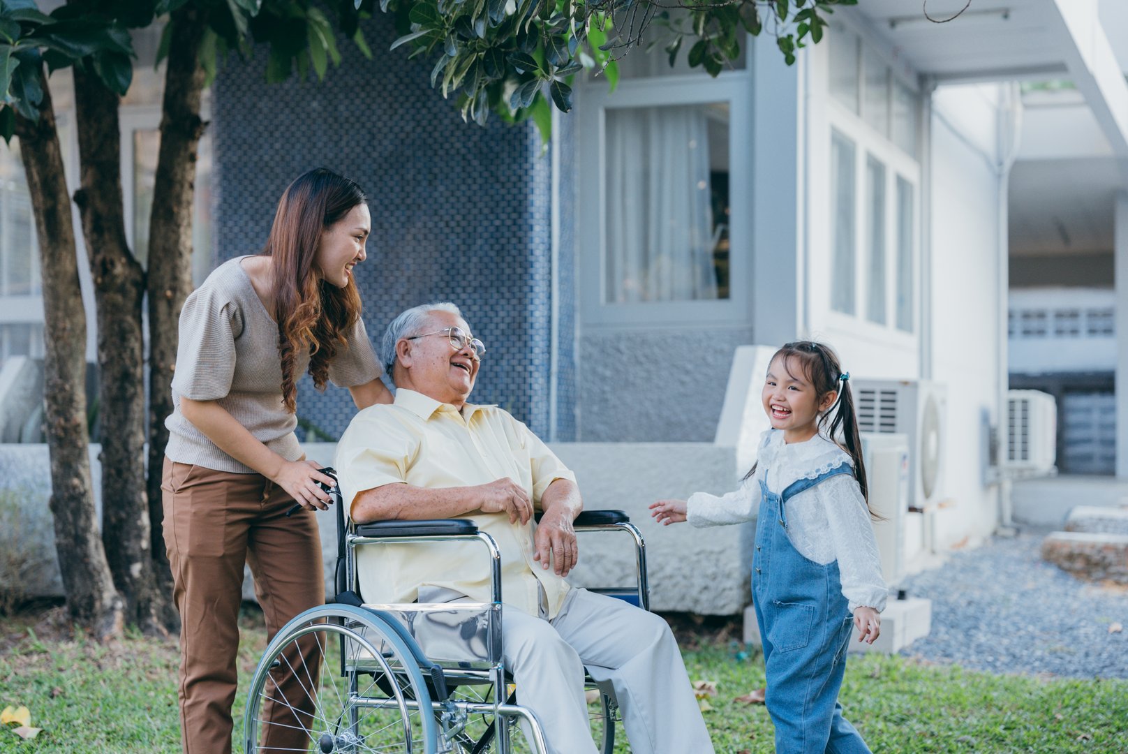 Disabled senior grandpa on wheelchair with grandchild and mother in park, Happy Asian multi generation family having fun together outdoors backyard, Grandpa elderly and little child smiling and laugh.