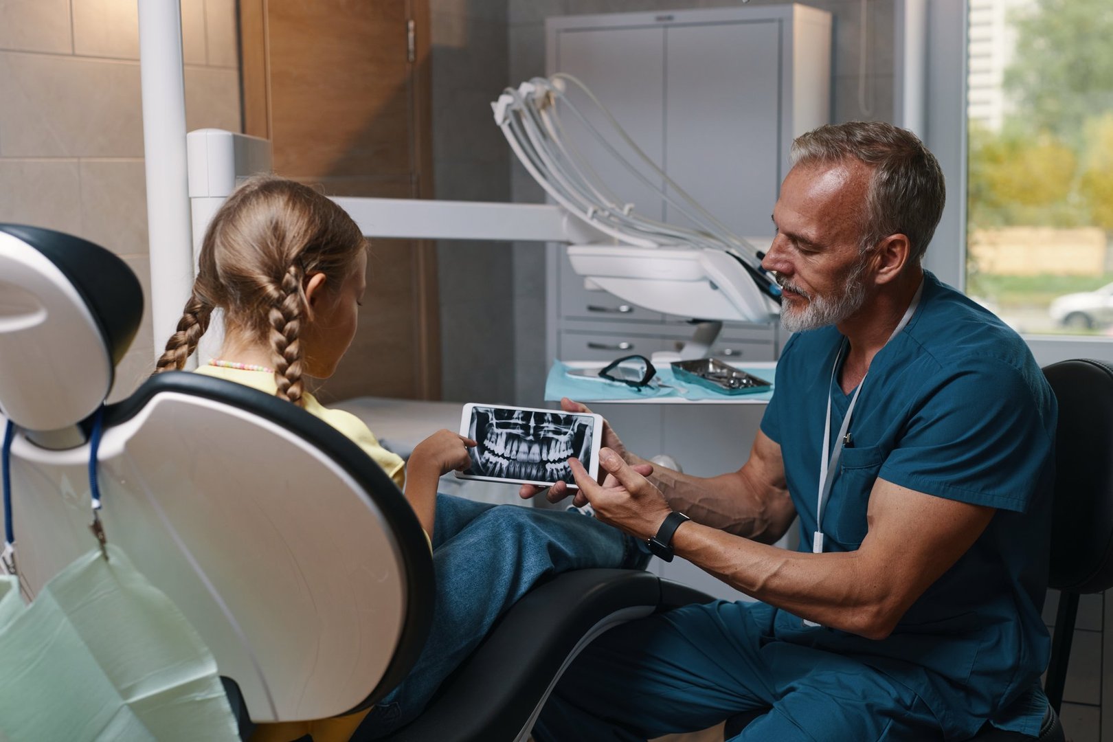 Dentist showing young patient dental X-rays on a tablet in modern dental clinic with equipment around. Dentist wearing scrubs and stethoscope, engaging young patient during consultation