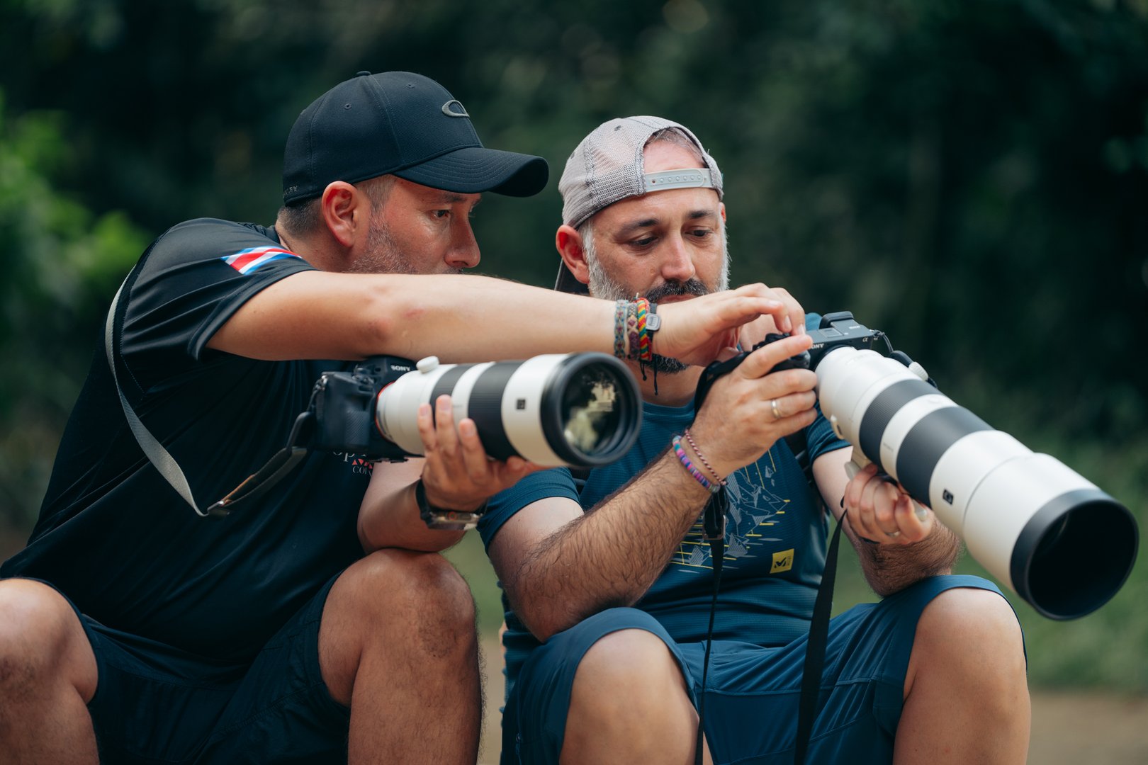 Two men sitting outdoors, examining a camera screen with large telephoto lenses attached, in a natural setting.