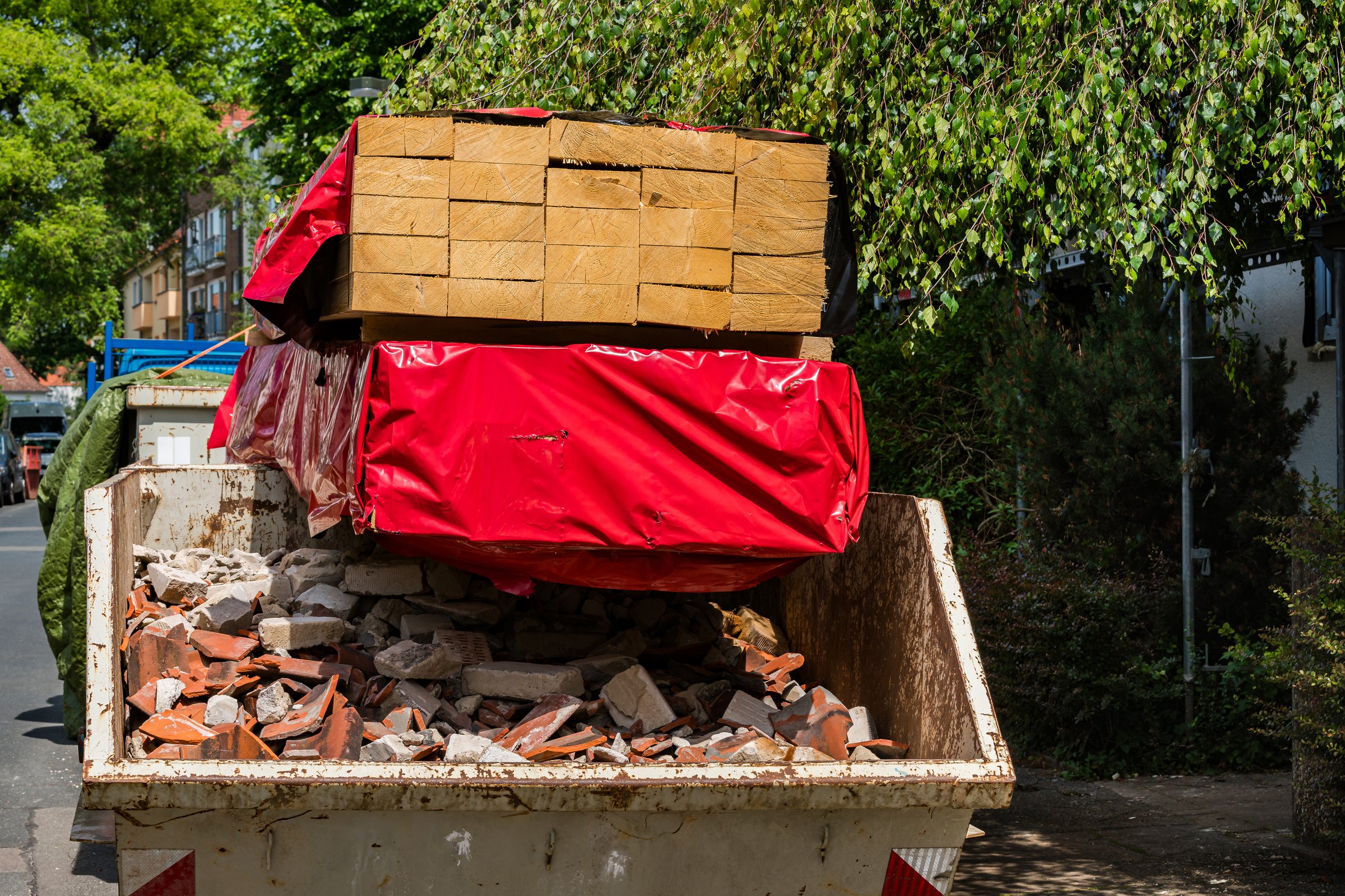 A large dumpster filled with bricks and construction materials sits on a city street, covered partially with a red tarp. Lush green trees provide shade in the background on a bright day.