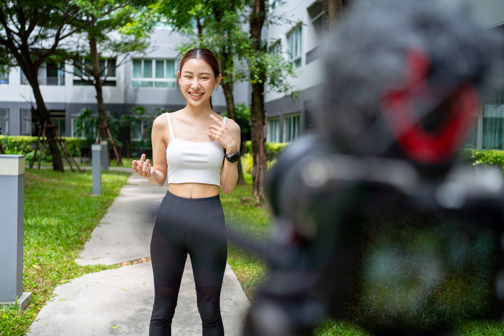 Cheerful young Asian woman vlogging outdoors, talking and gesturing to the camera for her video blog. Behind the scenes view of a female content creator presenting for her social media channel.Cheerful Asian fitness influencer recording a video for her social media channel in a park. Happy female vlogger flexing her bicep muscle for the camera, creating online content about health.