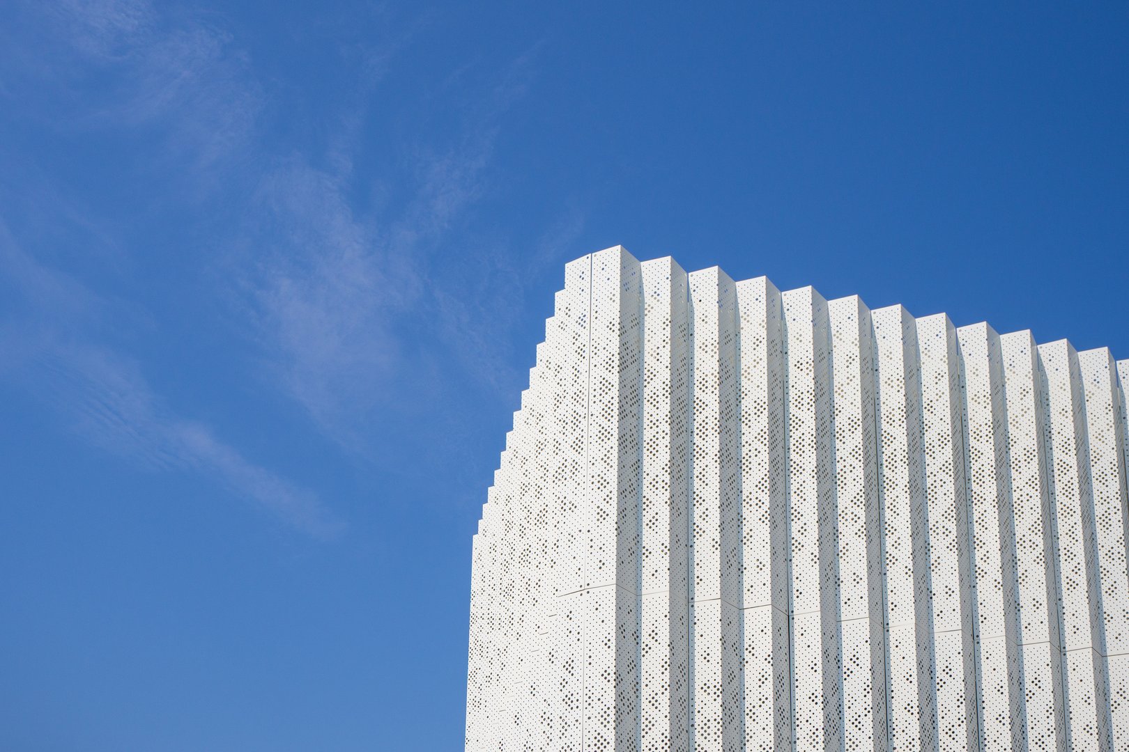 Decorative perforated metal facade panel. Modern exterior architectural element with vertical ribbed panels against blue sky background. White metal cladding with laser-cut pattern.