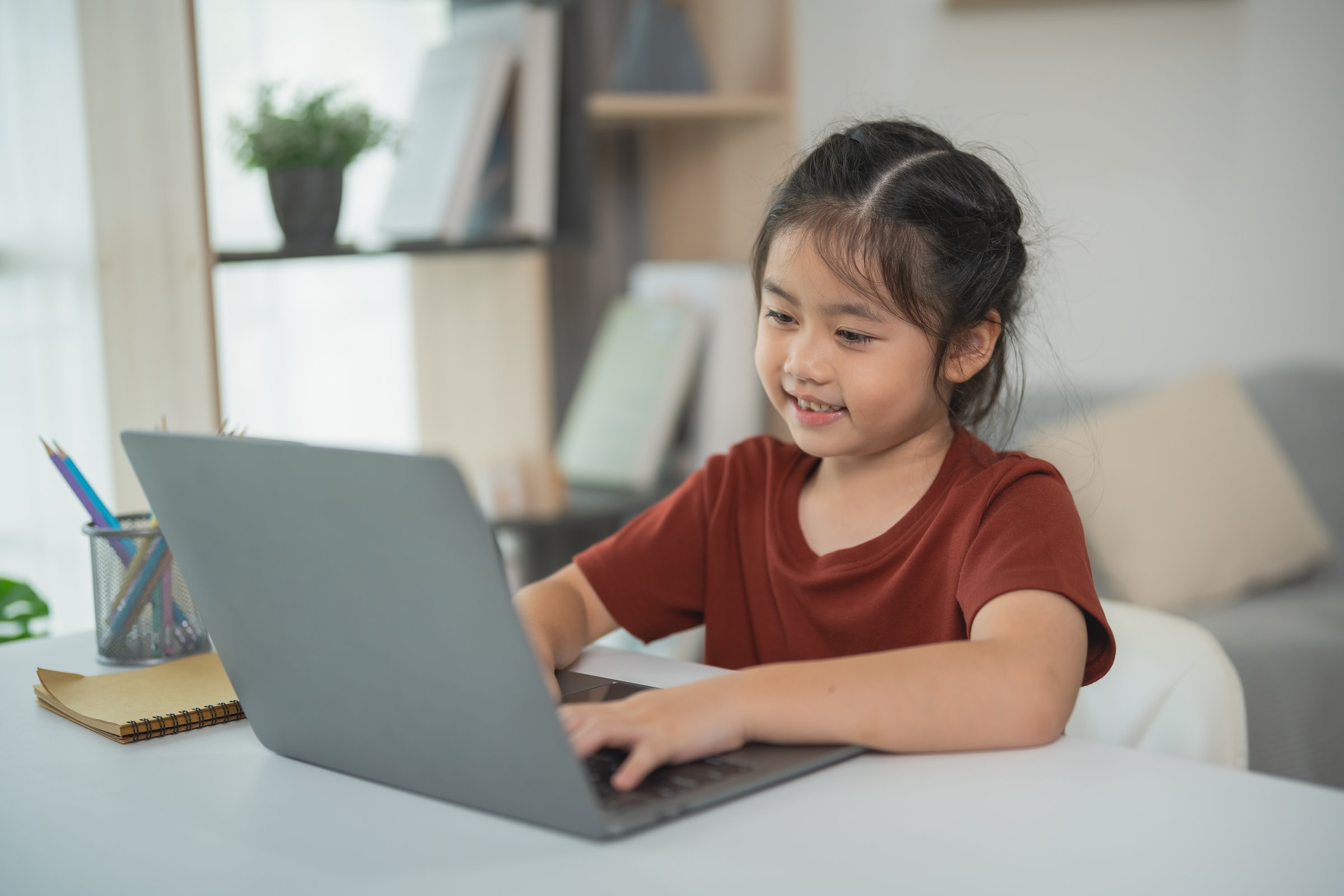 Young Girl Engaged in Online Learning, Smiling While Using Laptop at Home, Cozy Workspace with Books and Plants, Educational Environment for Kids