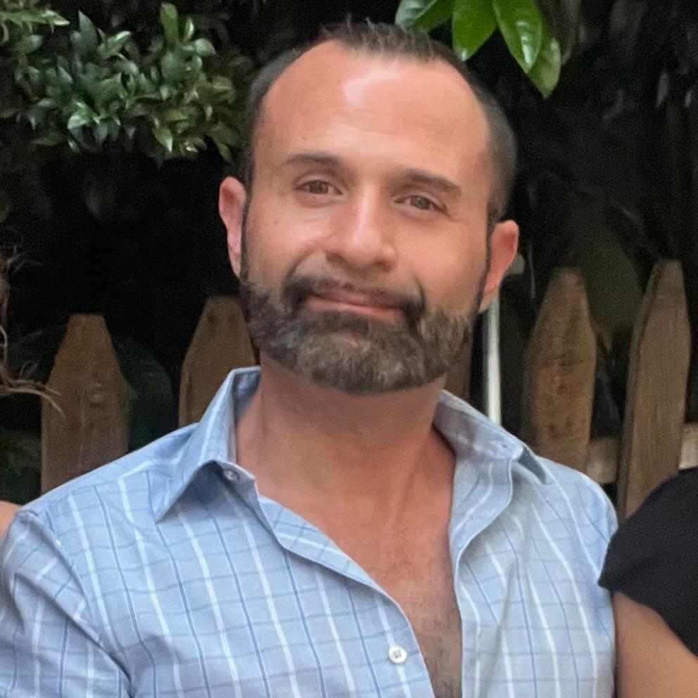 A man with a beard and a blue checkered shirt stands outdoors in front of a wooden fence and leafy background.