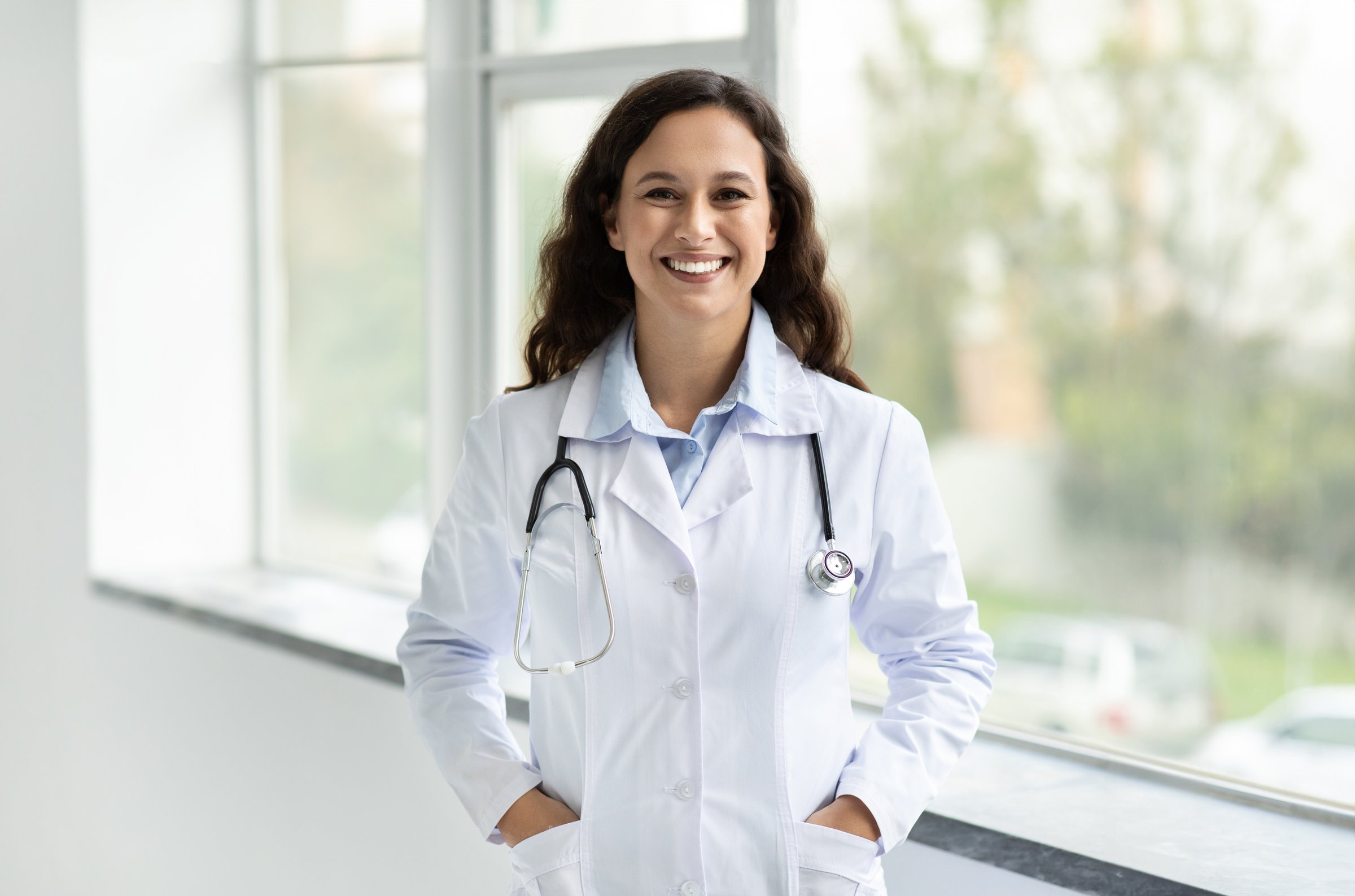 Healthcare Concept. Portrait of brunette european young woman doctor with stethoscope posing with hands in pockets over window background with free space at modern clinic, hospital